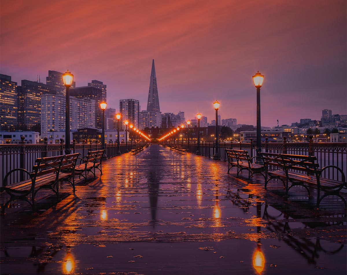 Blue hour view from Pier 7 in San Francisco with glowing street lamps and reflections leading toward the Transamerica Pyramid