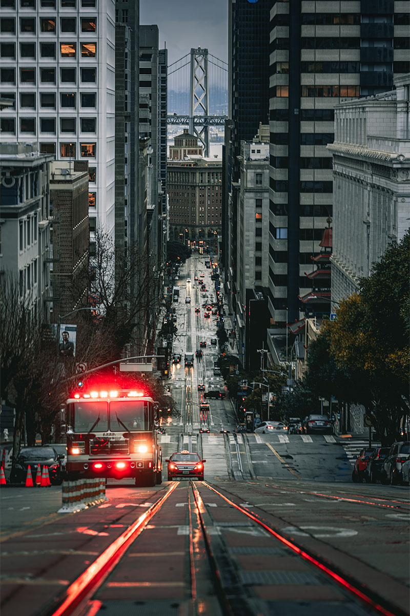 Fire truck and taxi on a rain-soaked San Francisco street with red lights reflected on wet pavement and the Bay Bridge in the distance