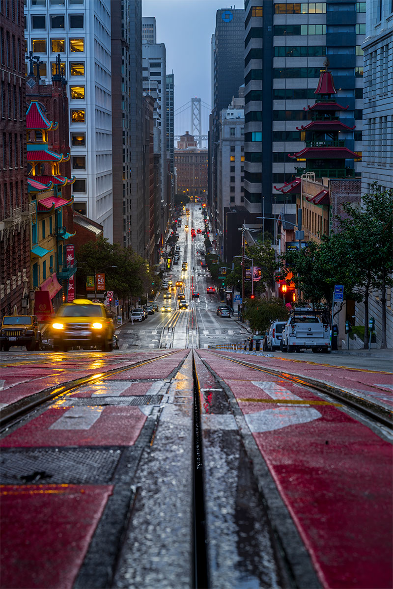 San Francisco street looking downhill along cable car tracks at blue hour with city lights, traffic, and the Bay Bridge in the distance